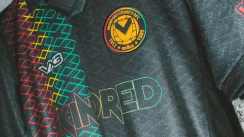 Newport County Close up shot of the black shirt, which features the stripes of the Caribbean colours in red, yellow and green and reads "Skindred" in their branding. The Newport County badge is yellow at the centre, with green, red and yellow around the rim.