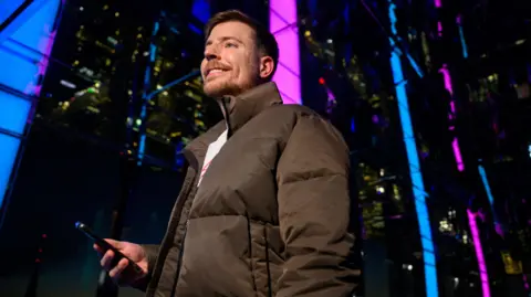 Getty Images MrBeast pictured outside in New York on a December evening in front of a glass building illuminated in blue and pink to promote Beast Games. He wears a brown puffer jacket and holds a smartphone in his hand.