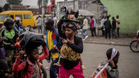 Getty Images A woman carries a sewing machine on her head and other belongings as she flees from Kibati, where fighting has intensified, towards the city of Goma on January 26, 2025.