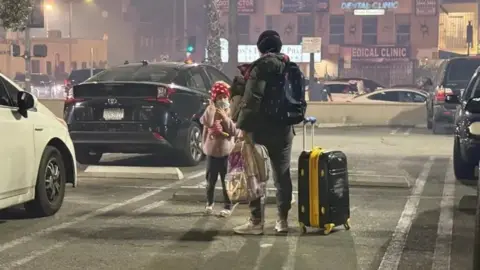 Christal Hayes A woman with her daughter stands on a road with a suitcase in one hand and a bag in another.