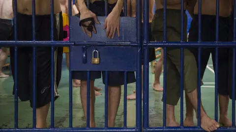 Getty Images Foreign detainees stand behind bars at an immigration detention centre in Bangkok.They are wearing dark-coloured shorts and pants, with some of their bare torsos and legs visible. One man's hands are also visble above the lock on the cell door.