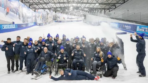 Royal British Legion A group of people all smiling at the camera at the bottom of an artificial indooer ski slope. Some people are in wheelchairs.