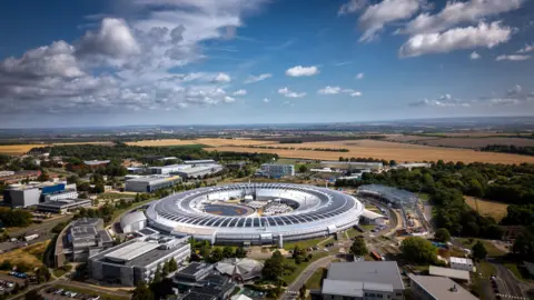 Tony Jolliffe/BBC Arial image of Diamond Light Source in Oxfordshire. The building is a large, silver donut-shaped ring which houses a particle accelerator inside. The countryside can be seen surrounding the machine.