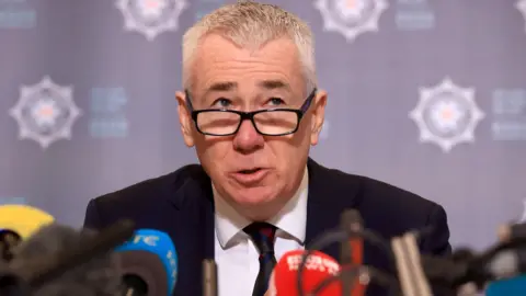 Getty Images Jon Boutcher, a man with grey, closely cropped hair and black-rimmed glasses, wearing a dark suit and tie, sitting in front of a large number of microphones with